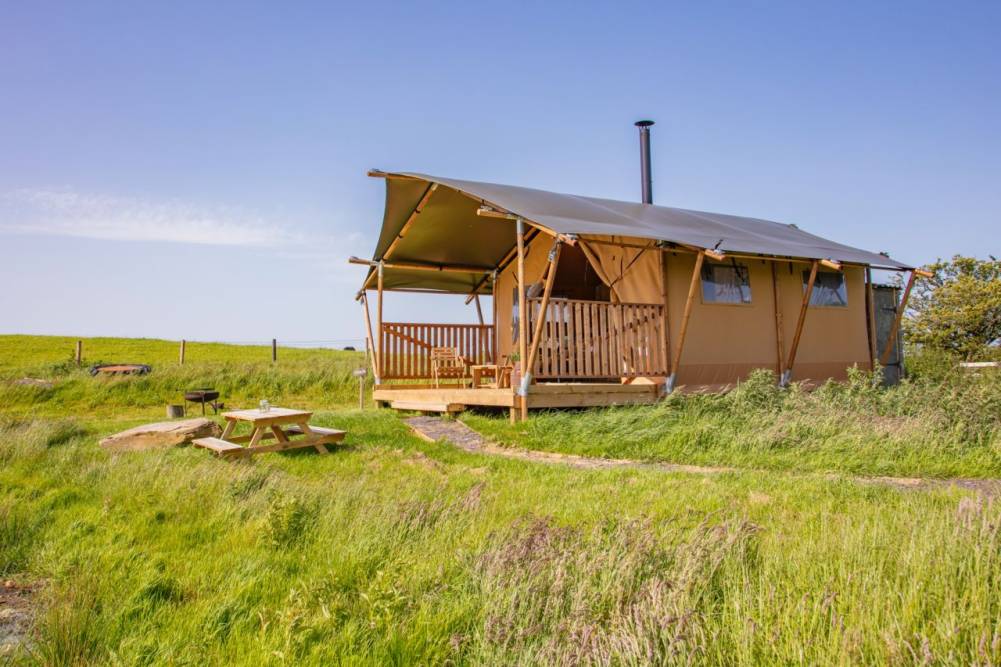 Canvas safari tent glamping lodge with a wooden deck, outdoor seating, and a picnic table surrounded by lush green meadows.