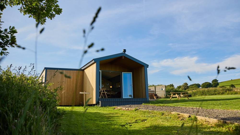 Gravel path leading up to the Dan-y-Dderwen wooden glamping pod with a picnic bench and hot tub on the lawn