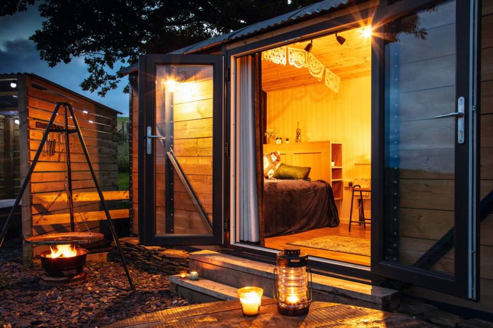 Cozy interior of a glamping cabin lit with warm lighting, viewed through open double doors with a lit fire pit and outdoor lantern in the foreground.