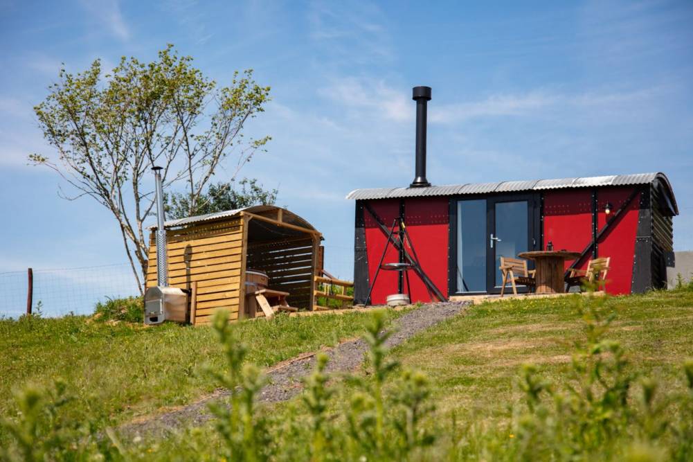 Red and black shepherd's hut with a metal roof, outdoor table, and a sheltered hot tub next to a tree in a grassy field
